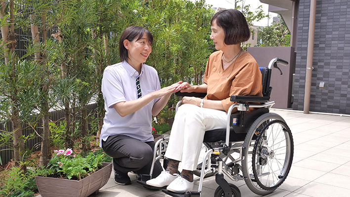車椅子の入居者に食事を提供するスタッフ。屋外テラスでの日常ケアの風景。